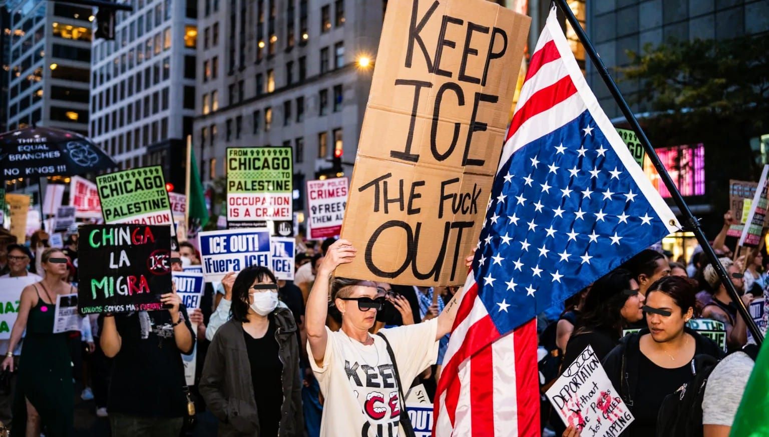Against a background of tall buildings, protesters march and hold anti-ICE/anti-occupation signs along with an upside down US flag