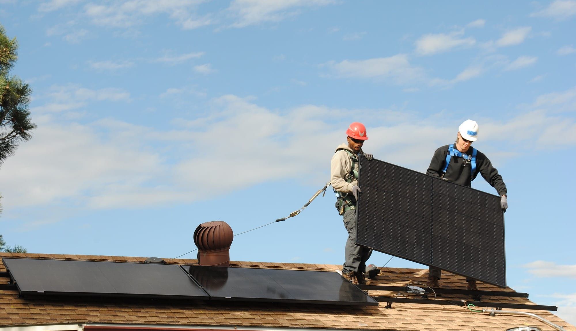 Two people on a roof in construction hardhats and safety harnesses install a solar panel next to two others, against a bright blue sky.