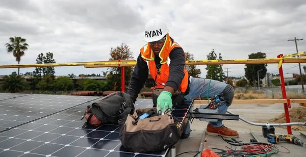A young Black man in a helmet that says "RYAN" and other safety gear works on a solar installation, with a fence, cloudy sky and trees in background.