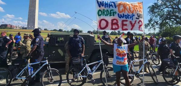 With Washington Monument in the background, federalized cops and militarized vehicles are staged with the Washington Monument in the background as a middle aged Black woman protests with a c