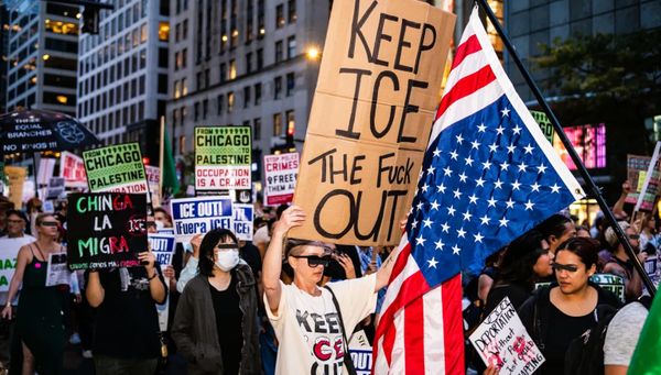 Against a background of tall buildings, protesters march and hold anti-ICE/anti-occupation signs along with an upside down US flag