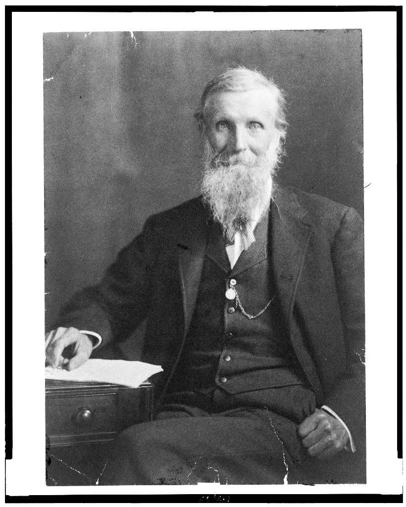 old man with long gray beard sitting next to a table with papers beneath his hand