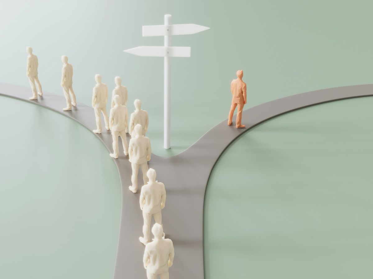 Plastic human figures on a gray plastic road that diverges. Most figurines take the path on the left. The tan-colored figurine takes the path to the right.
