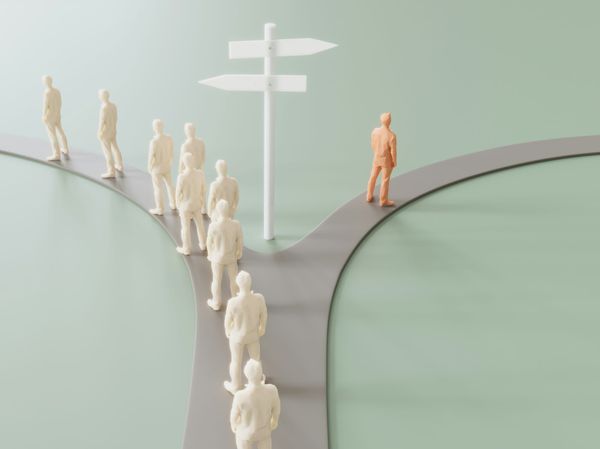 Plastic human figures on a gray plastic road that diverges. Most figurines take the path on the left. The tan-colored figurine takes the path to the right.