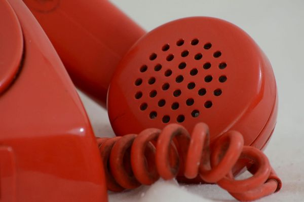 Photo of a red rotary phone, showing only one corner of the phone’s square-shaped body, a section of the coiled cord, and the mouthpiece end of the receiver.