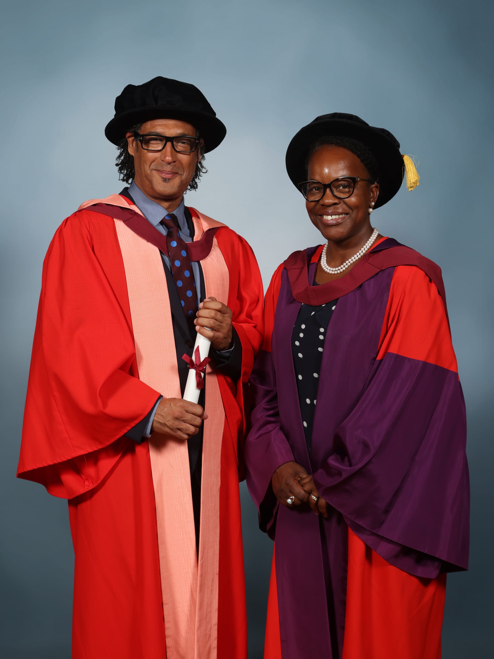 Olusoga stands in front of a grey background, alongside Dr. Gournet. Olusoga is wearing red academic robes over a suit and tie, and a black hat with gold tassel. Gournet is wearing an identical hat, with similar robes (except with a purple accent dominating the centre) over a black blouse with white spots. She also wears a pearl necklace.