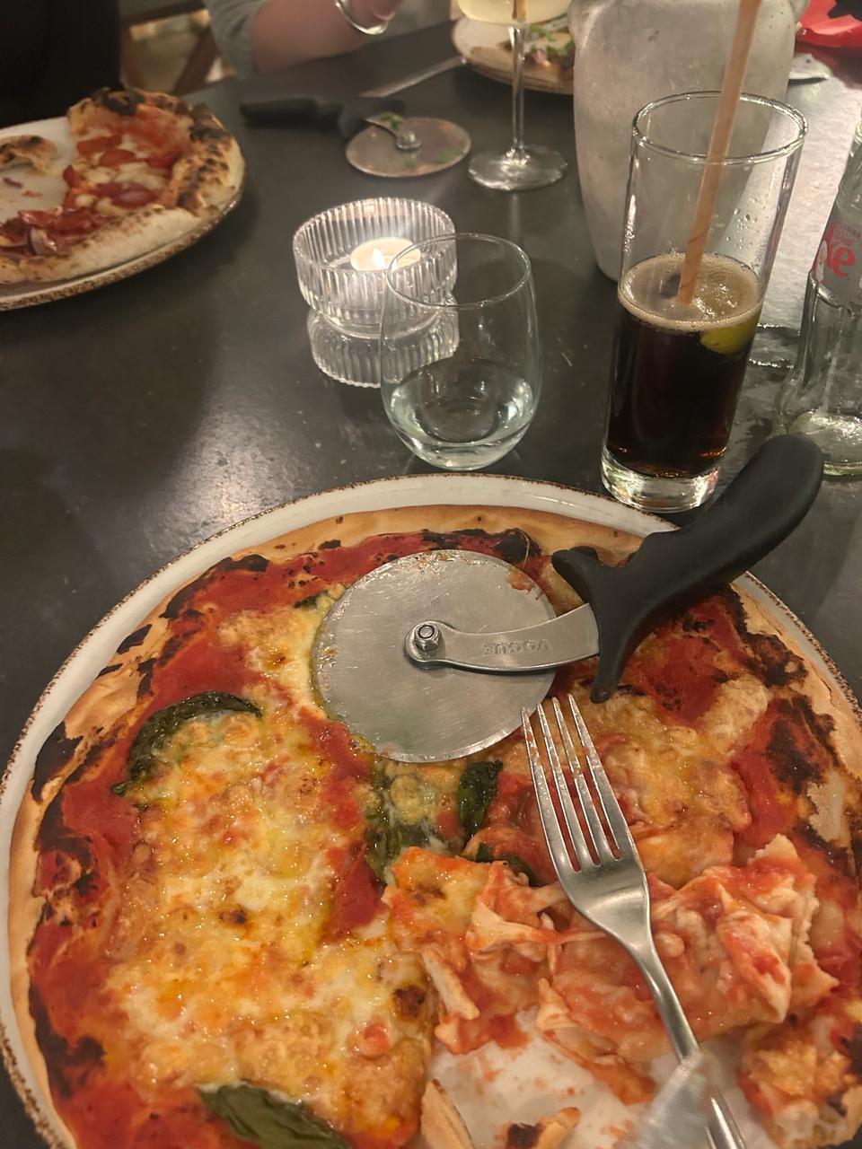 A pizza with a fork and pizza cutter on top of it. Drinks and someone else's pizza in the background.
