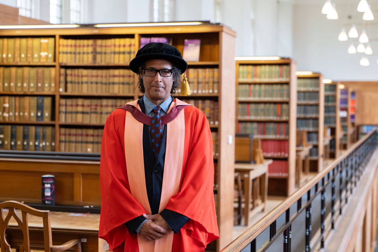 Olusoga stands in front of a desk and bookcase in the Wills Memorial Library, alongside the balcony railings. He is wearing red academic robes, and a black hat with gold tassel.