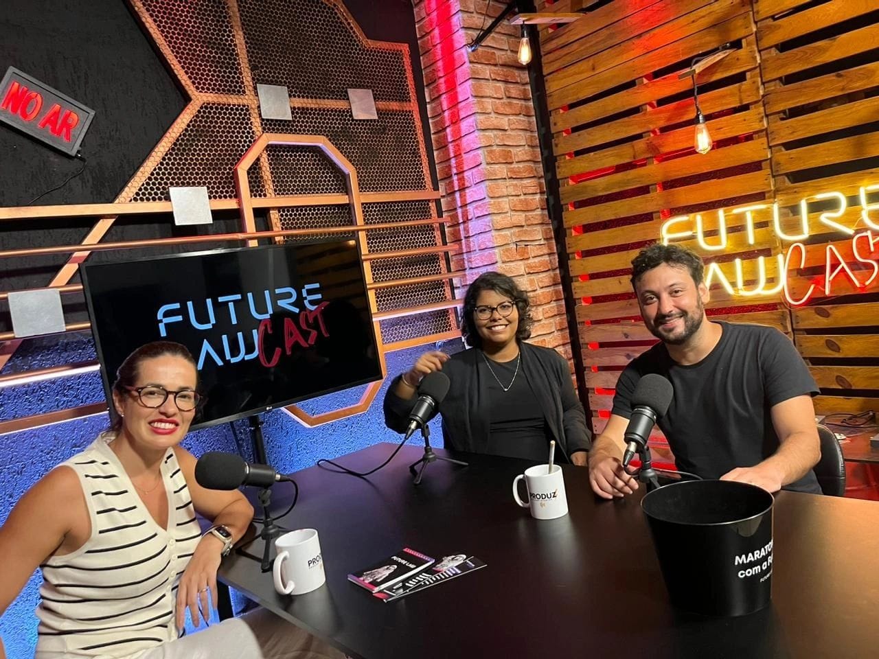 Na imagem: três integrantes se reúnem ao redor de uma mesa gravando o podcast
