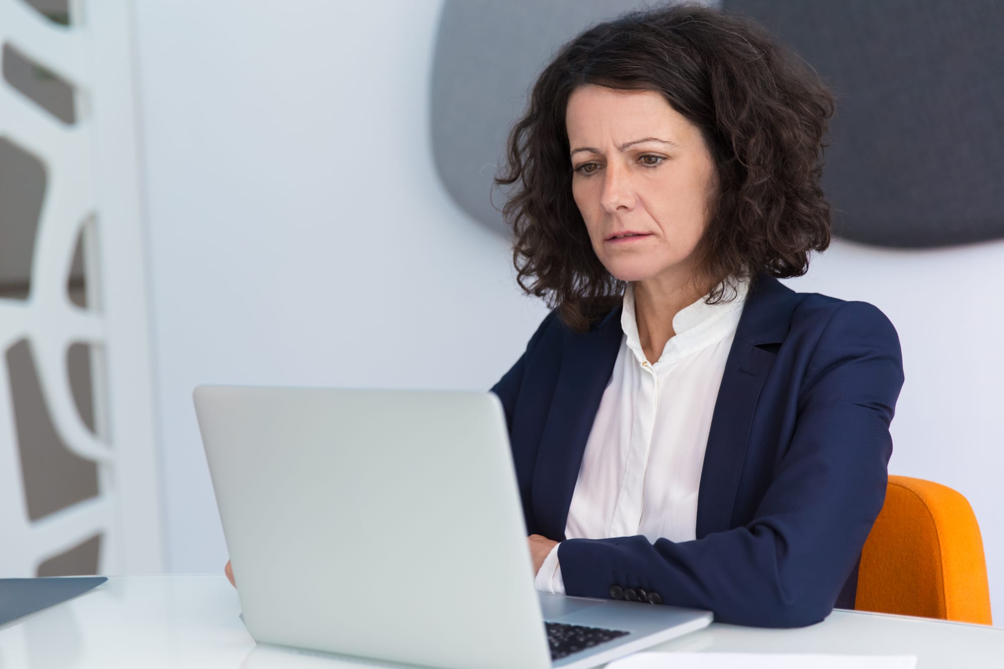 Na imagem: mulher séria vestida de blazer e camisa social observa atentamente a tela do notebook