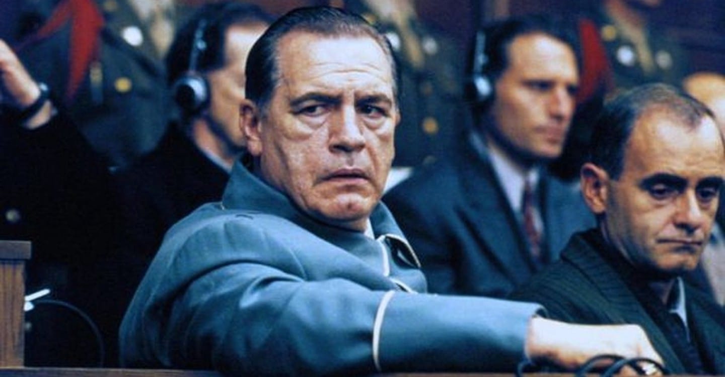 Na imagem: o ator Brian Cox, na pele de Hermman, está sendo na mesa dos réus olhando para o lado com tom de preocupação.