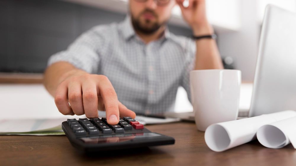 Na imagem: homem está em mesa do escritório clicando em uma calculadora com uma caneca ao lado. 