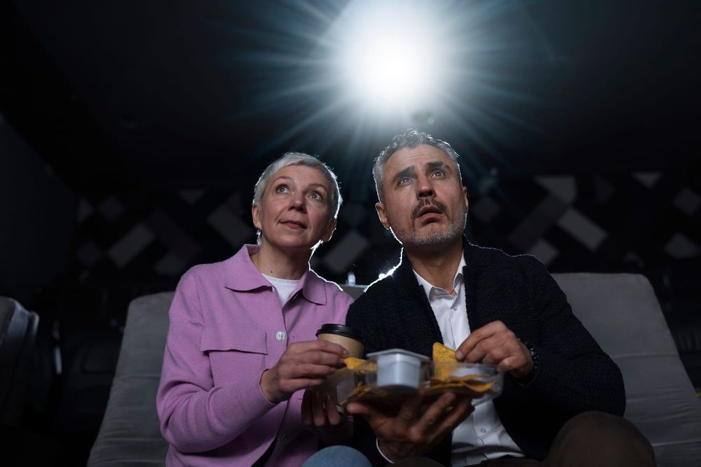 Na foto: um casal come lanches em suas poltronas em frente a um projetor de cinema