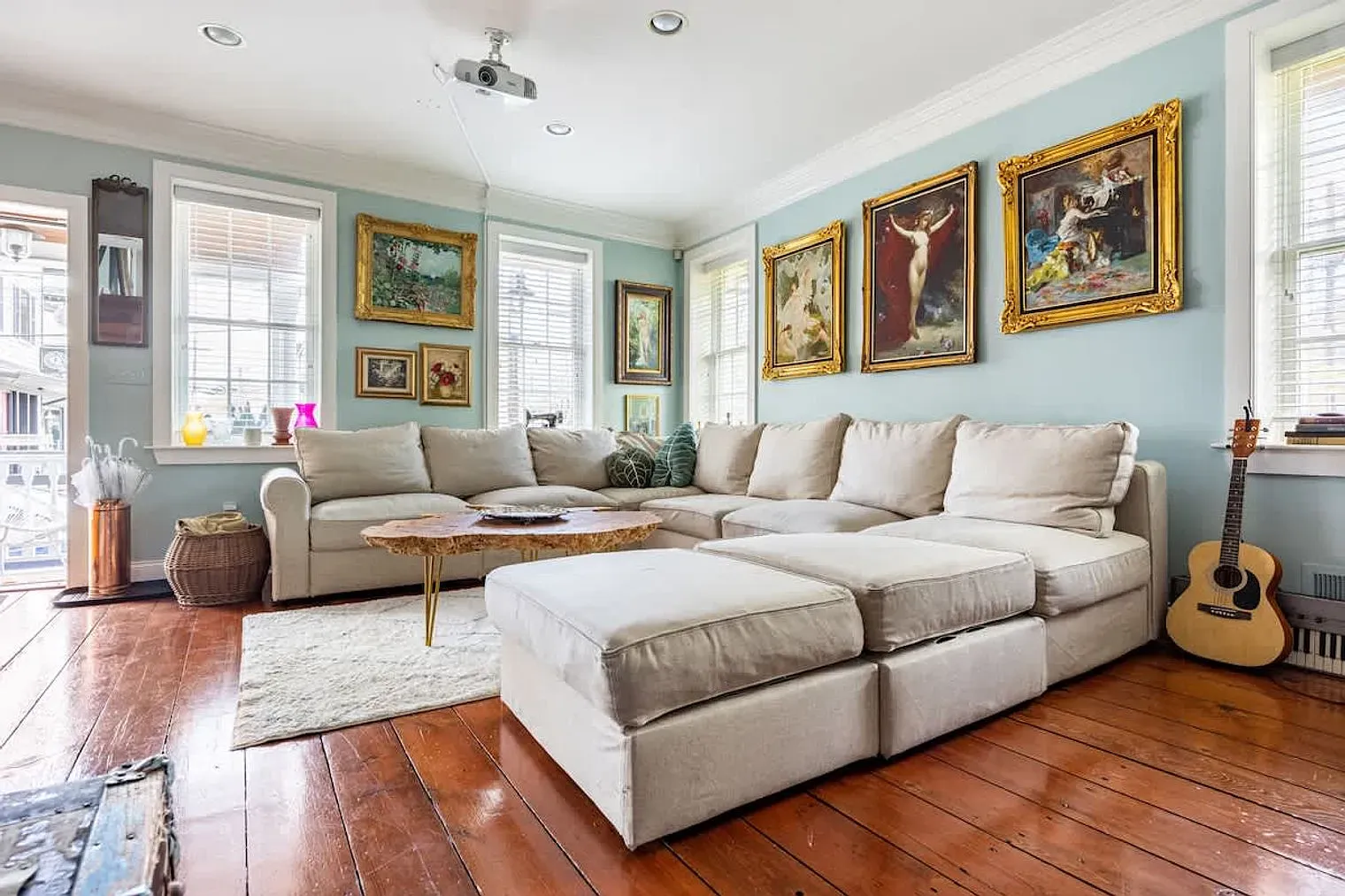 a large couch fills the room. paintings hang on the wall and an acoustic guitar leans against the wall. large windows fill the room with light