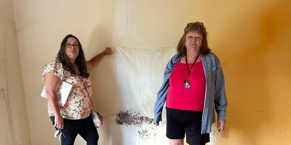 two women stand by a rust-stained wall with mold growing on it