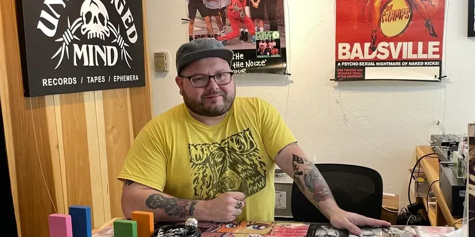 a smiling tattooed man standing at the check out counter of his record store
