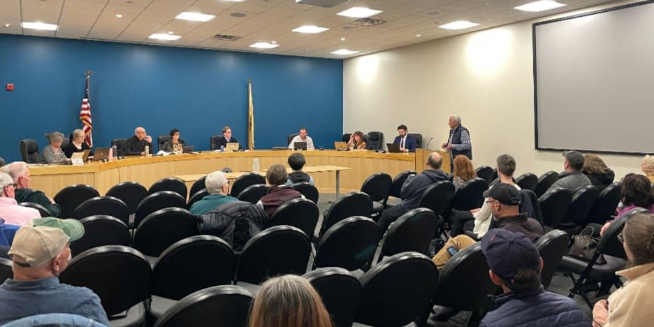a man speaks to the city council while other citizens watch and listen
