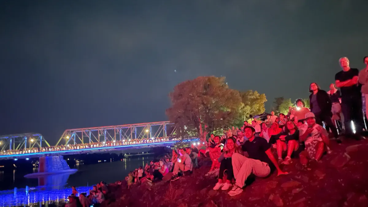 a crowd of people sitting along the banks of the Delaware River looking up at the fireworks