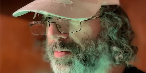 a bearded man with long, curly hair, wearing glasses and a baseball cap