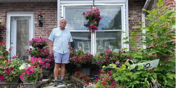 a white-haired man stands on his front steps, surrounded by flowers