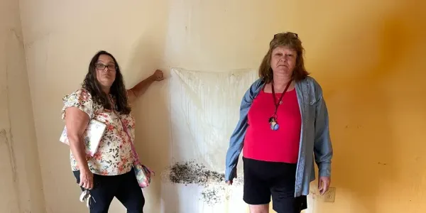 two women stand by a rust-stained wall with mold growing on it