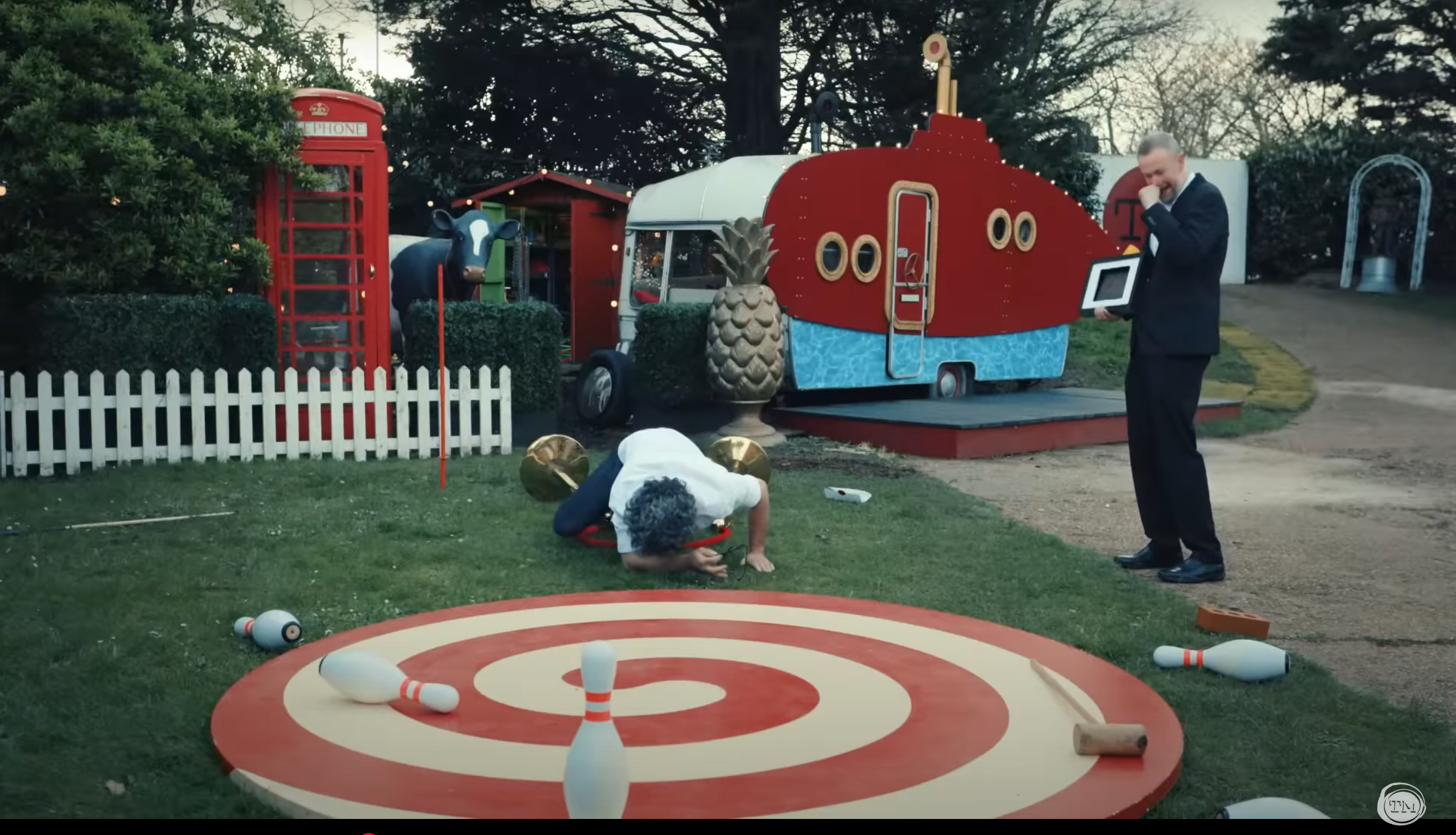 Jason Mantzoukas faceplants on the Taskmaster grounds while Little Alex Horne holds his fist to his mouth to suppress a giggle.