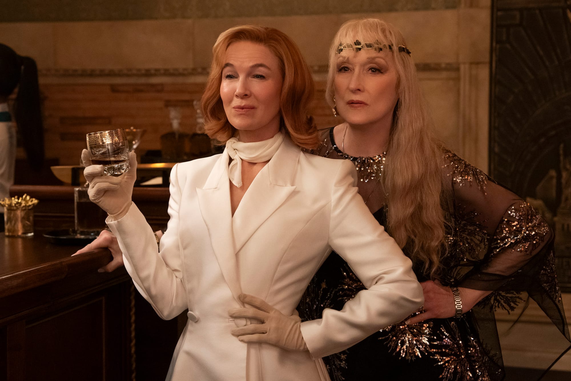 Renée Zellweger wears an all-white outfit and holds a rocks glass while standing in front of Meryl Streep.