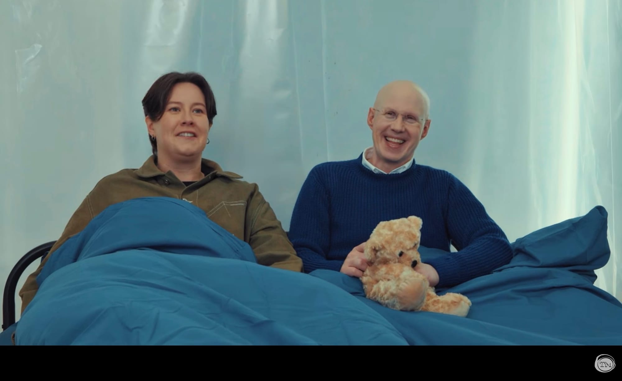 Chloe Petts and Matt Lucas in a bed. Lucas holds a teddy bear.