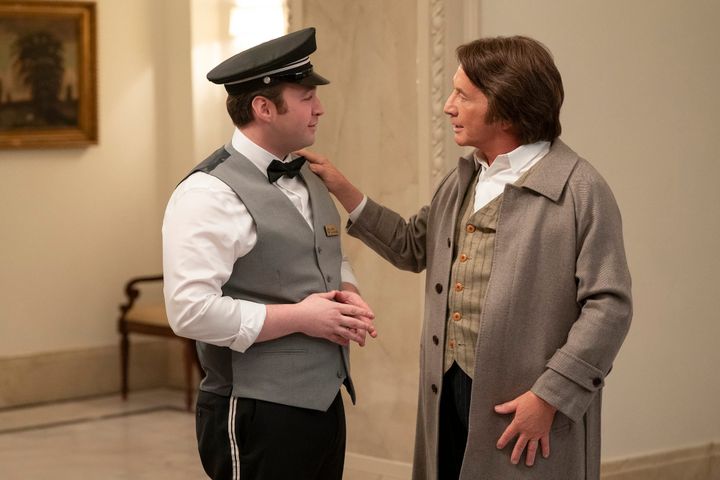 A man with brown hair wearing a gray vest, black bow tie, and black cap speaks with a man with long brown hair, a gray overcoat, and pinstriped vest.