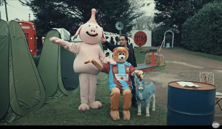 A pig mascot with a chess pawn on its head, a bear wearing a football jersey, a small plastic donkey, and Sanjeev Bhaskar, all on the Taskmaster grounds.