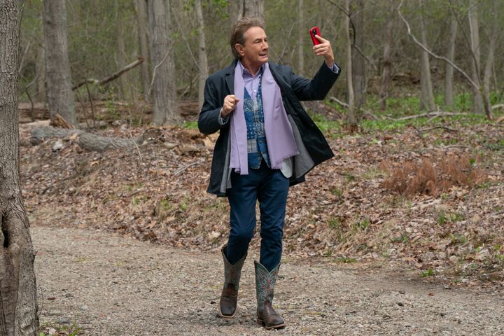 Martin Short is in the woods, wearing a blue blazer, purple scarf, patchwork vest, and gaudy cowboy boots while looking at a smartphone in a red case.