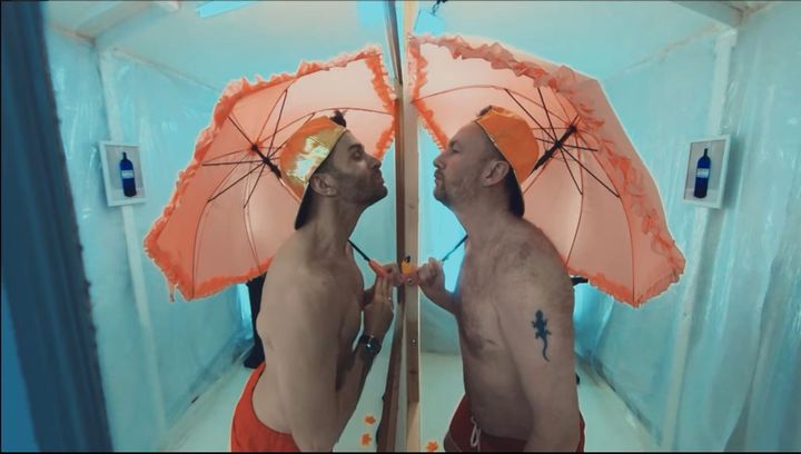 Joel Dommett and Alex Horne, both topless, face each other while carrying a frilly parasol.