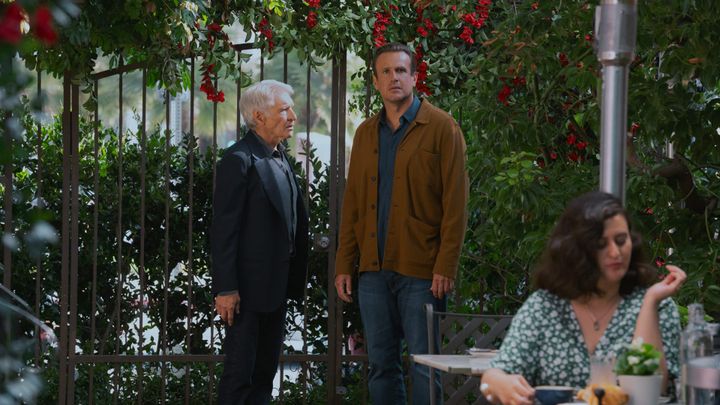 Harrison Ford and Jason Segel stand in front of a gate and some flowers at an outdoor restaurant