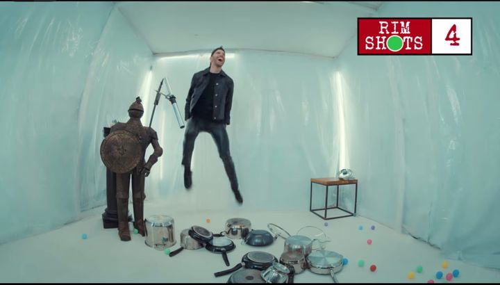 Joel Dommett leaps into the air amid a room full of pots, pans, and colored balls.