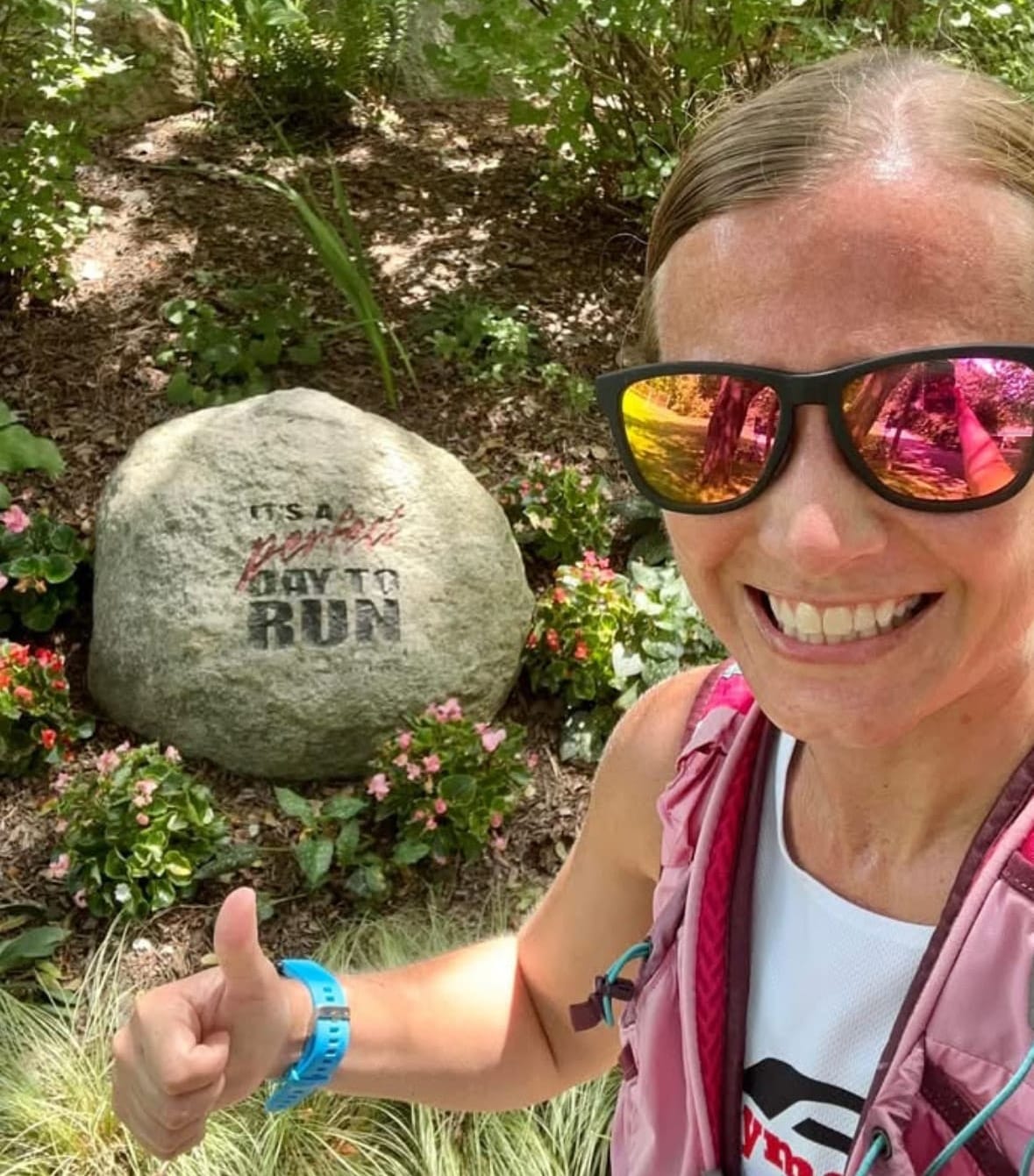 A woman wearing sunglasses and giving a thumbs up taking a selfie outside with a big rock in the background with words painted on it that read "it's a perfect day to run"