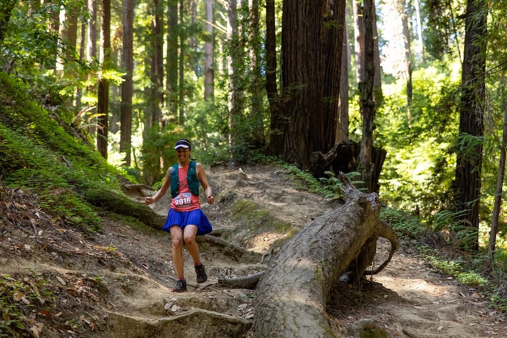 A woman in a blue running skirt and hydration vest running down a dirt trail with large trees in the background
