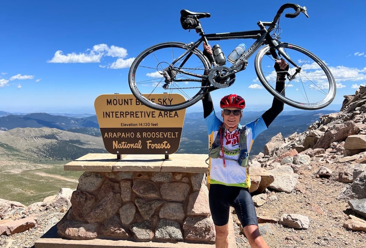 A cyclist holding his bike above his head at the summit of a mountain with blue skies in the background