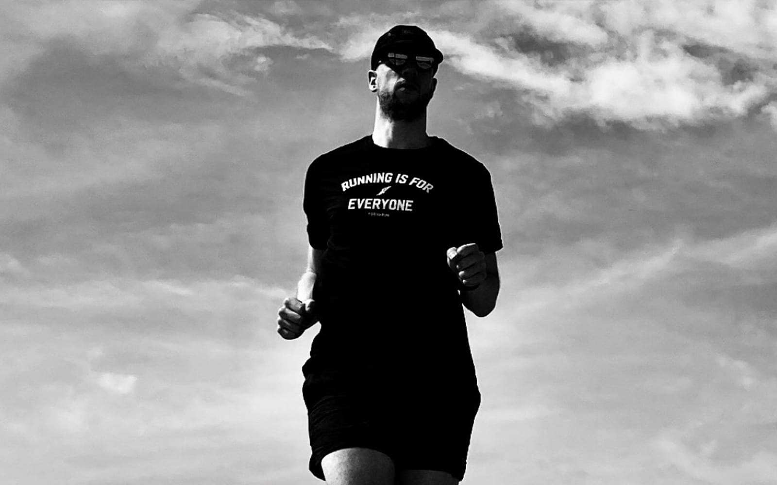 A man running, wearing black shorts, t-shirt, hat, and sunglasses