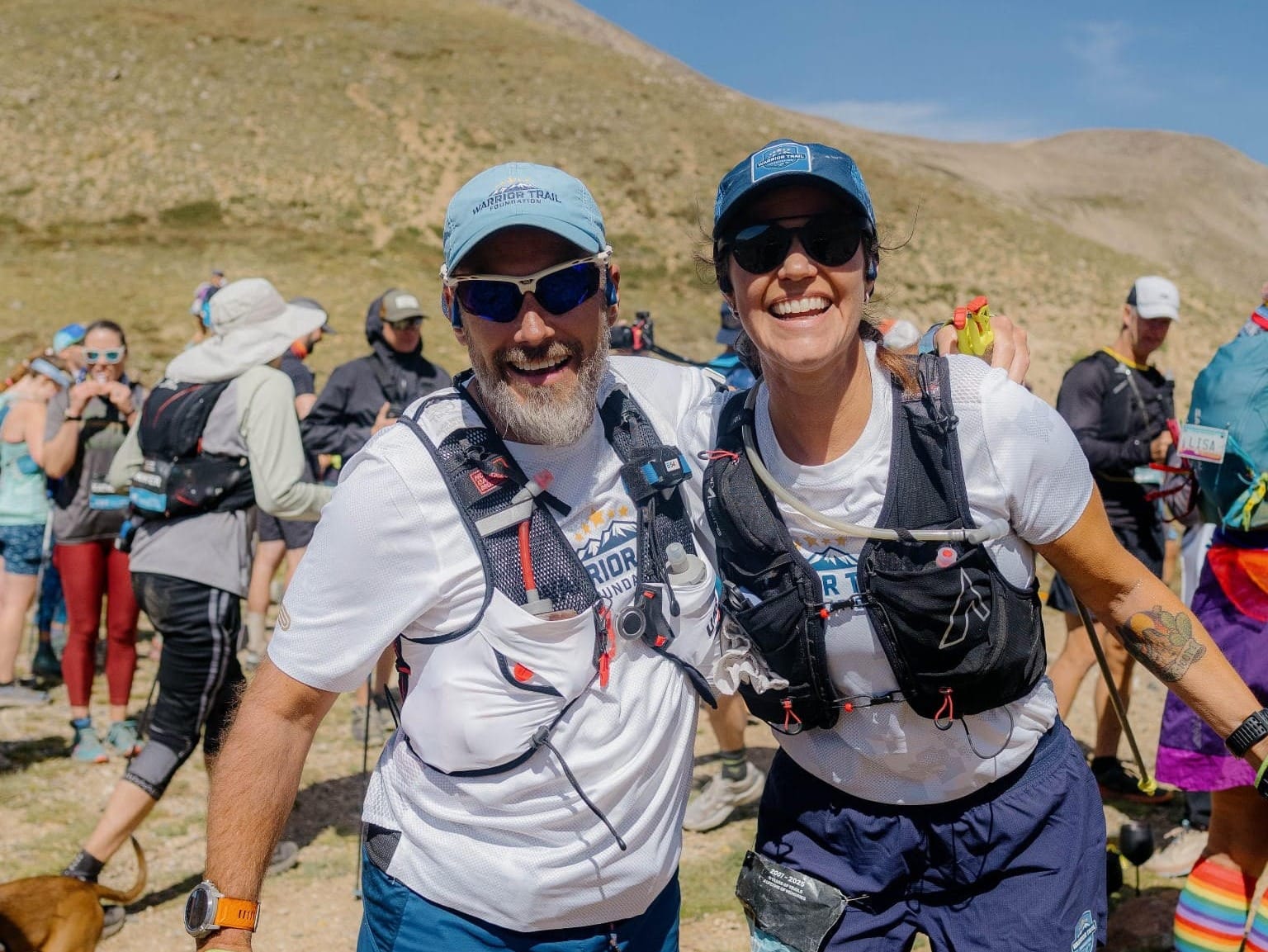 A man and a woman in trail running gear, smiling and posing together outside, with mountains and other runners in the background