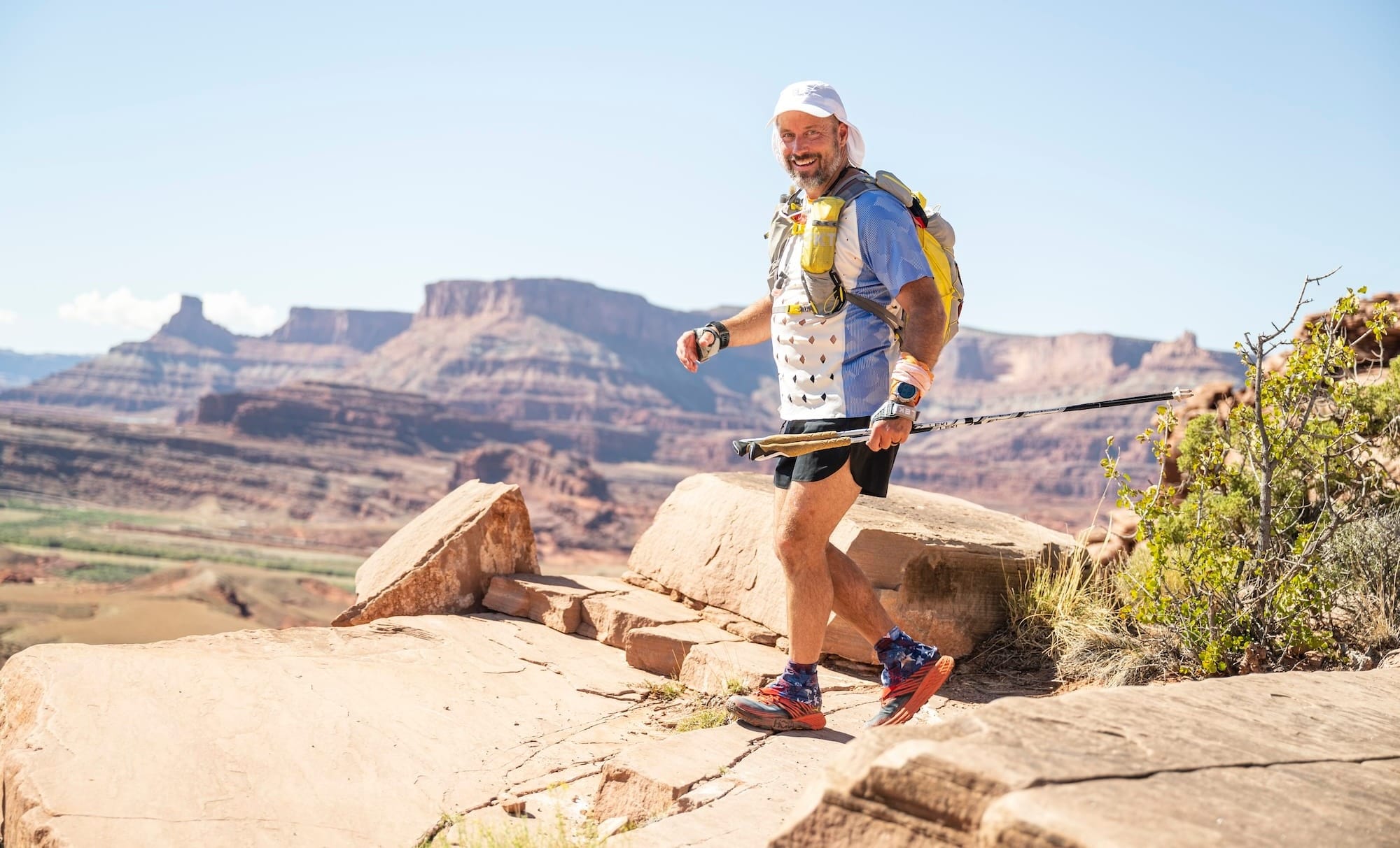 A man in trail running gear, smiling while walking across rocky terrain with mountains in the background