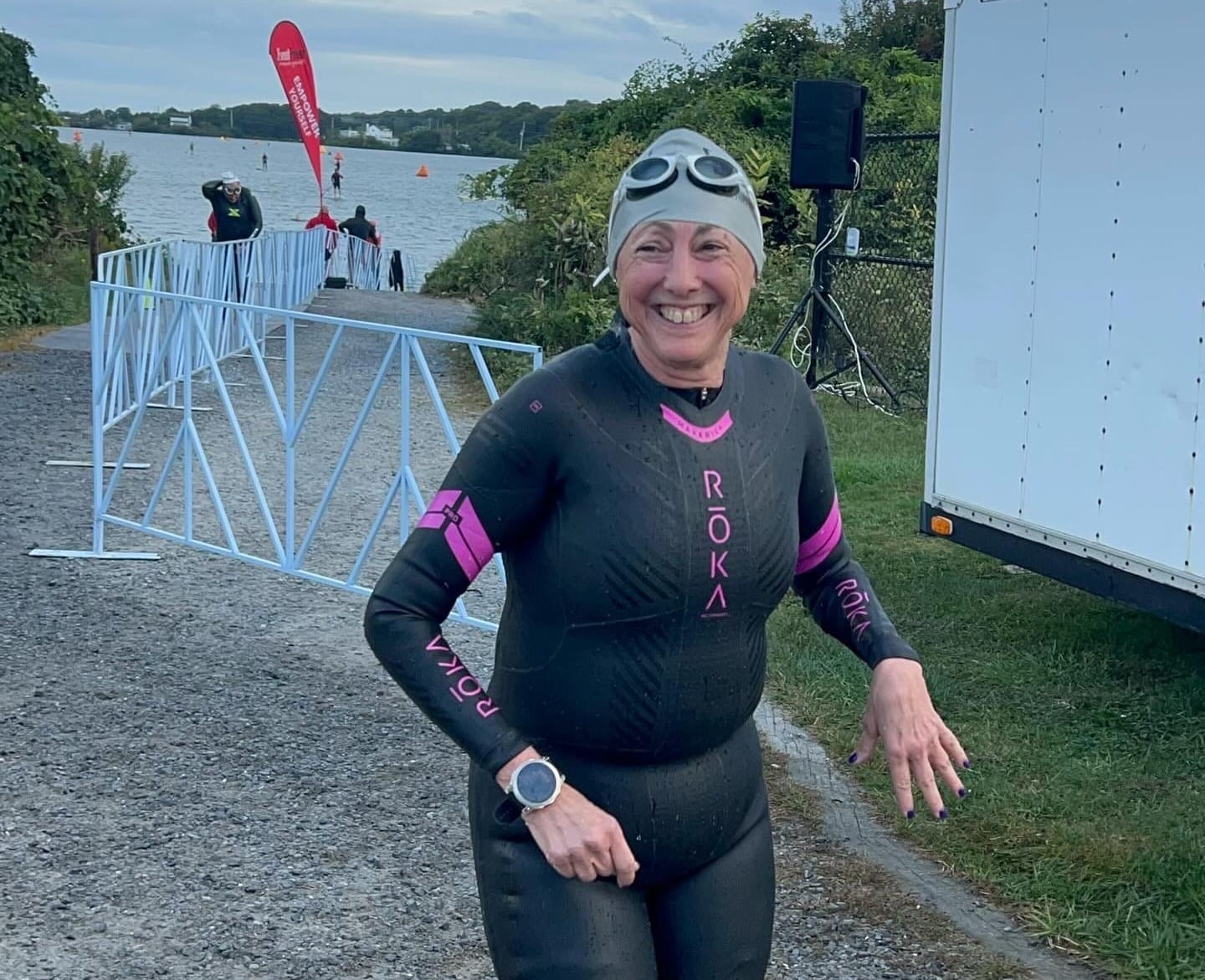 A woman smiling, wearing a wetsuit, swim cap, and goggles, with a lake in the background