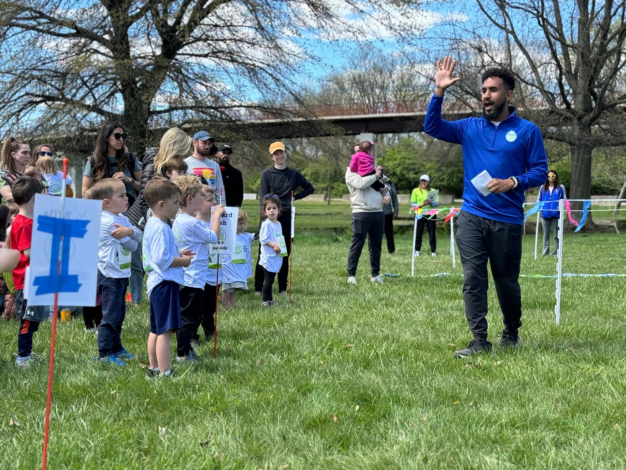 A man raising his hand and talking to a group of children standing together on the grass with parents in the background