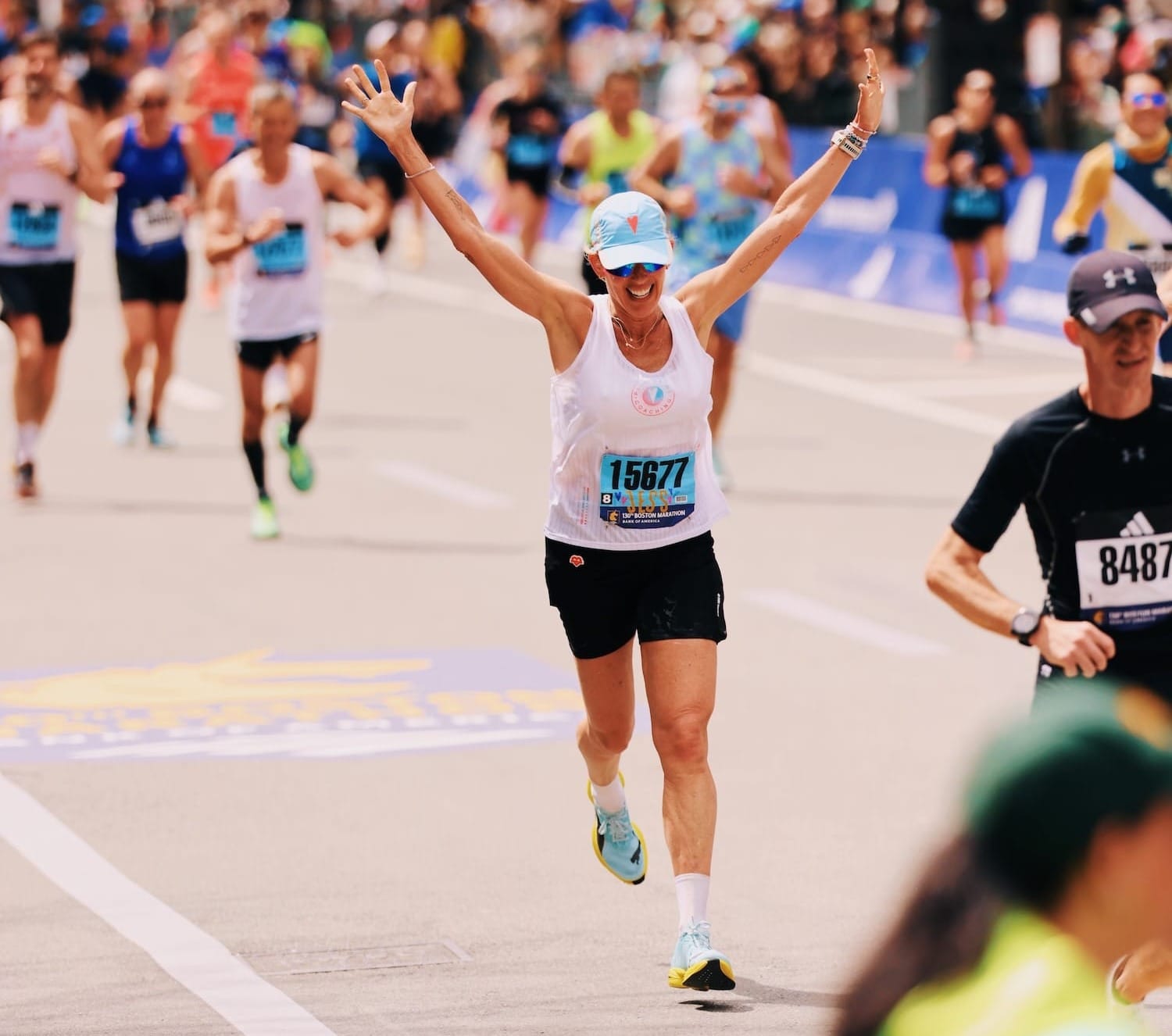 A woman running a road race, smiling and with her arms raised, with other runners around her