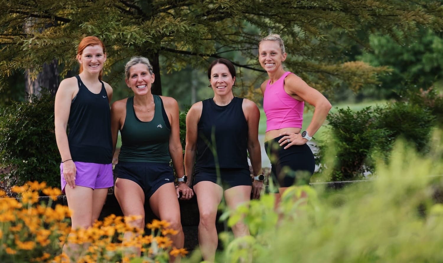 Four women in athletic apparel standing together with trees, flowers, and greenery around them