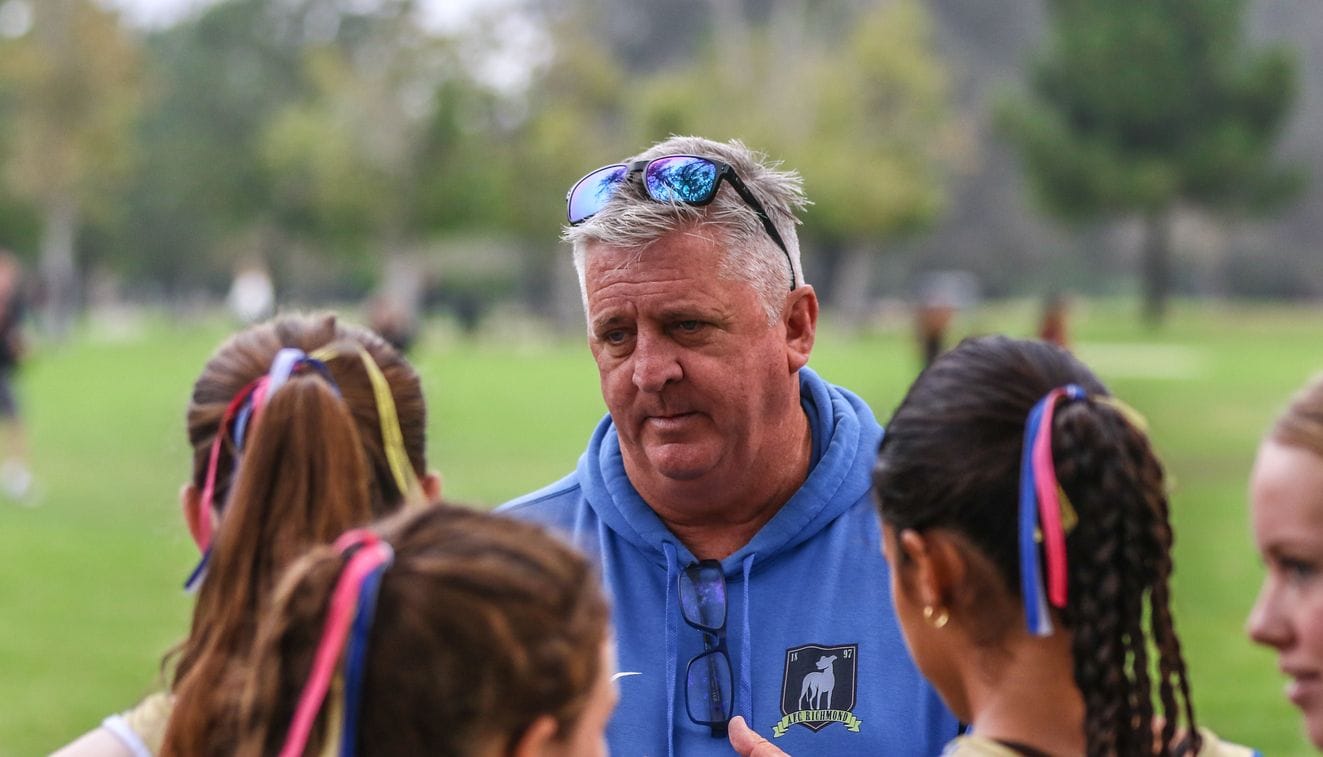 A coach talking to several female athletes outside with blurred grass and trees in the background