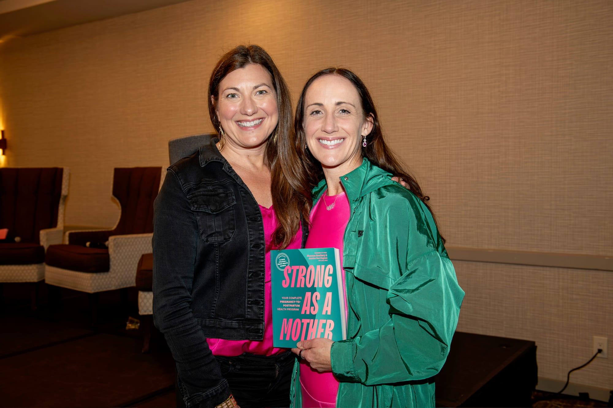 Two woman posing together inside a room while one holds a book in front of them.