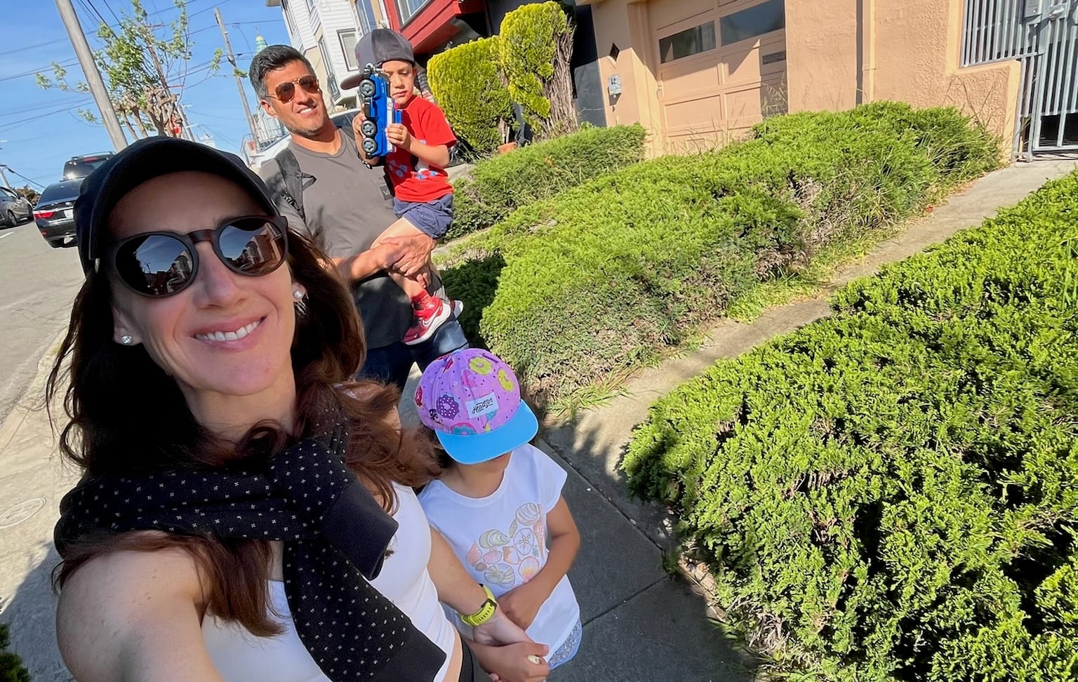 A women walking and taking a selfie outside with a man and two kids walking behind her