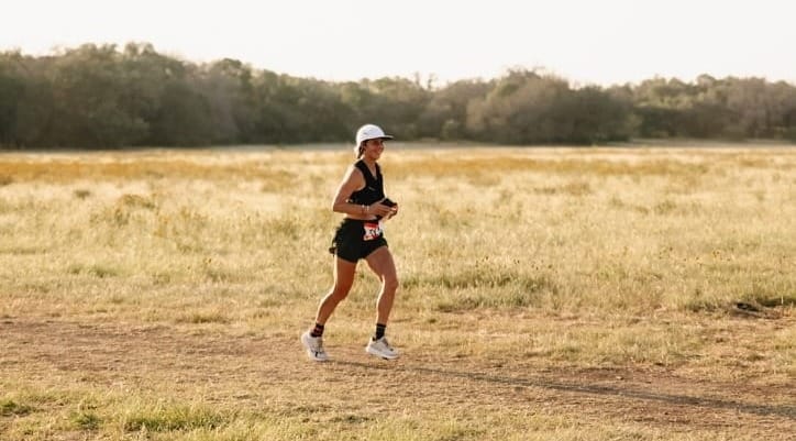 A woman running on a trail surrounded by dry grass with trees in the background