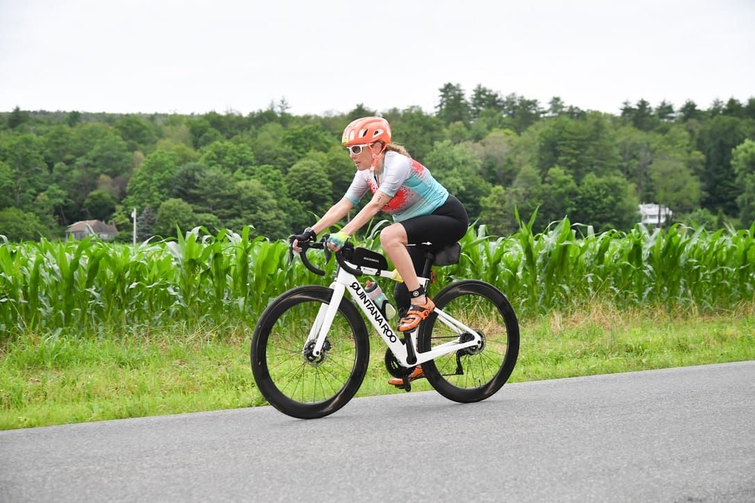 A woman riding a triathlon bike on a road with green trees and a corn field in the background
