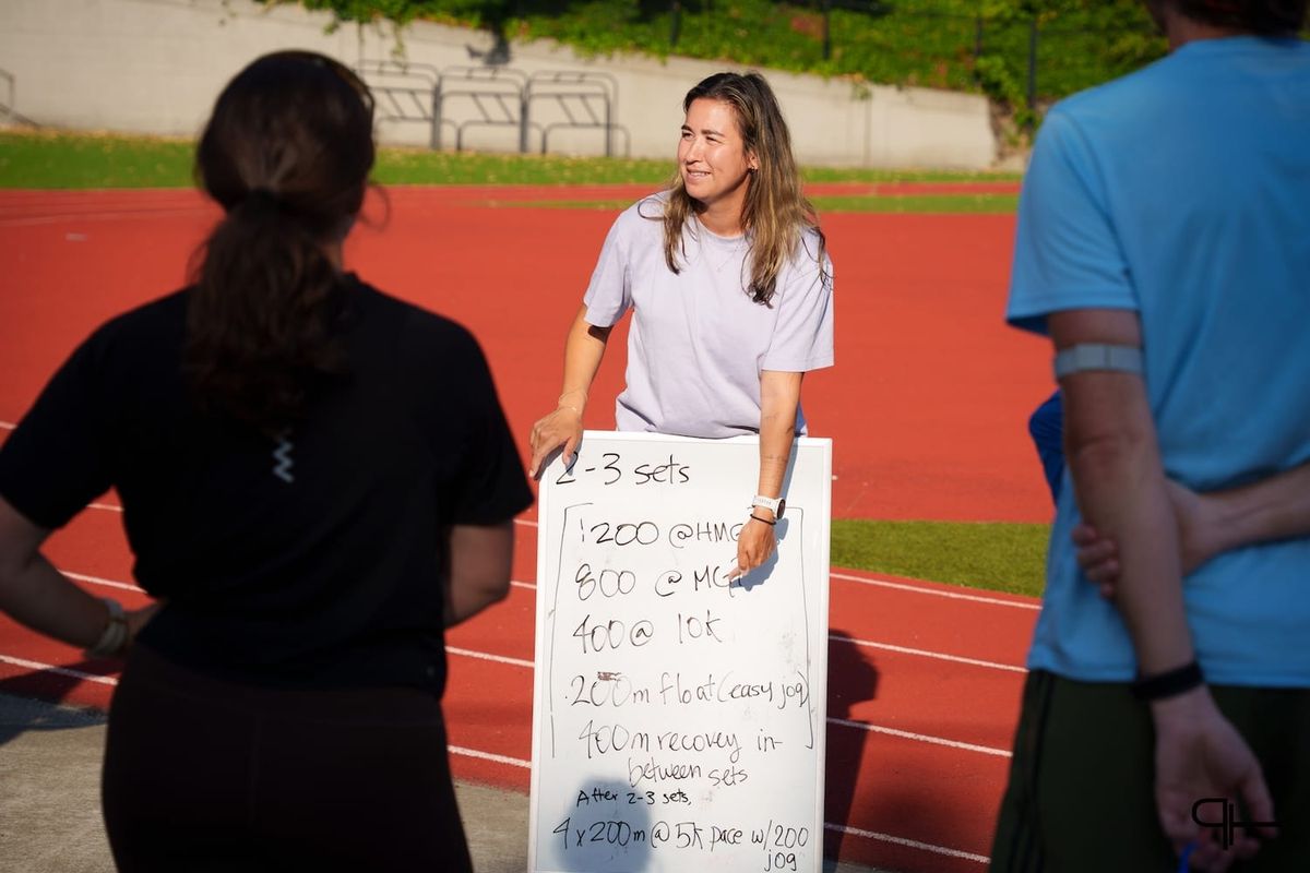 A women holding a whiteboard with a workout written on it while standing outside on a track